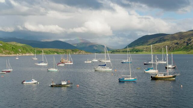 High-to-low drone shot over Ullapool harbour reveals fishing and leisure boats beneath a dramatic sky, with a rainbow stretching down Loch Broom toward distant Highland peaks.