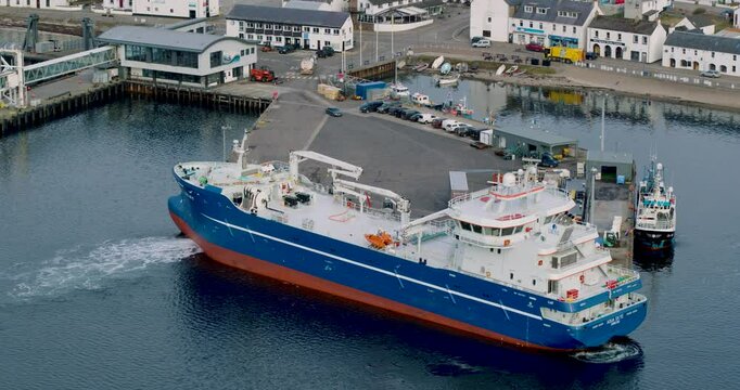 Drone shot of fishing carrier ship AQUA SKYE arriving into Ullapool on a crisp winter day, fully loaded, with snow-covered mountains in the background and calm loch waters.