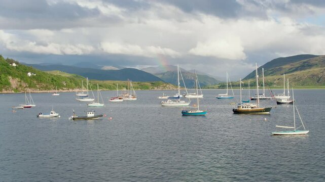 Rolling left-to-right drone shot over Ullapool harbour captures fishing and leisure boats below a dramatic sky, with a rainbow stretching down Loch Broom toward the Highland peaks.