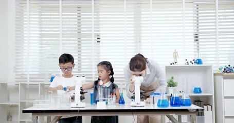Asian teacher using microscope to examine science sample while Asian boy and girl aged about 6 prepare glassware beside her during engaging elementary school classroom experiment - Powered by Adobe