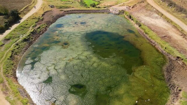 An aerial drone shot captures a high-altitude pond near Raqch'i, Peru, with the stunning snow-capped Andes mountains visible in the background of the rural landscape and two tourists on mountain road