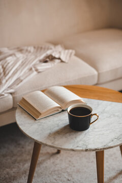 Mug of black fresh coffee with open paper book staying on marble table and velour couch in living room close up. Evening time. Relaxation.