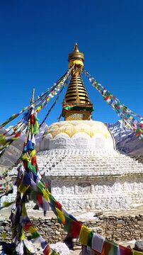 transparent buddhist stupa, golden spire, vibrant lungta prayer flags billow on rocky terrain before majestic snow-capped himalayan peaks under azure sky, crane video emphasizing grandeur.