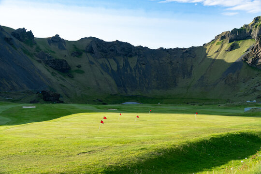 View of a vibrant green golf course nestled within a dramatic volcanic crater, red flags punctuating the landscape, under a clear blue sky, VestmannaeyjabÃ¦r, VestmannaeyjabÃ¦r, Iceland.