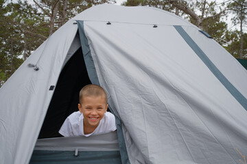 A happy boy with a wide smile looks out from the opening of a gray camping tent, ready for outdoor...