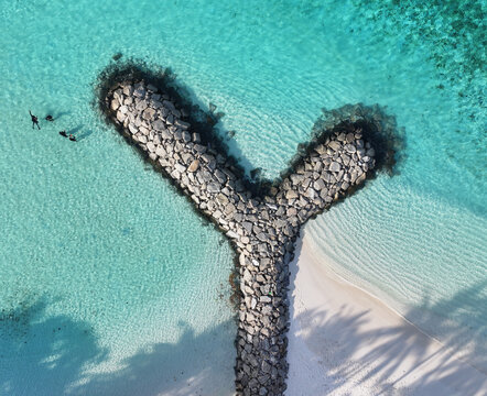 Aerial view of a rocky pier splitting azure waters meeting white sands under palm shadows, Kendhoo, Baa Atoll, Maldives.