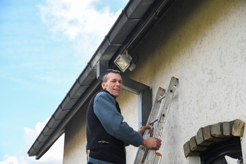 Professional electrician installing powerful automatic LED light with motion sensor under house roof, standing on ladder with outside of residential building, looking at camera