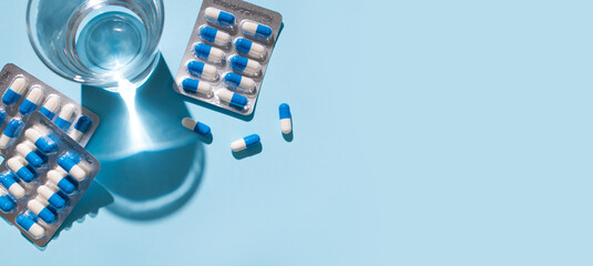 Health and medicine concept. White and blue capsules in silver blister pack, glass of water on blue background. Tablets, pills, vitamins, food supplements, probiotic. Pharmacology, health and beauty 