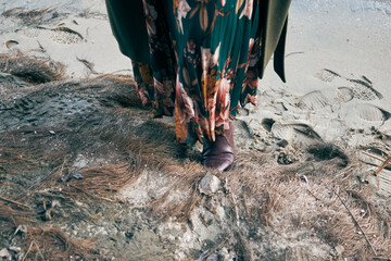 Woman in floral dress and boots standing on sandy beach with dry grass