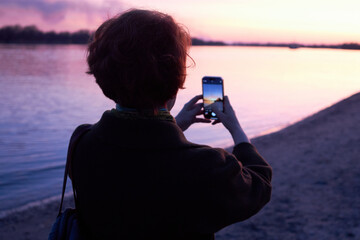 Woman photographing sunset by the river with smartphone on a tranquil evening
