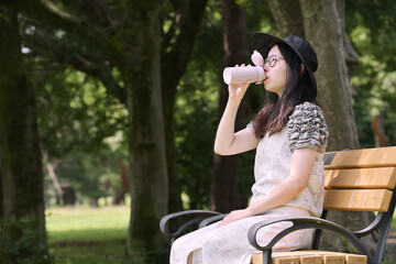木陰のベンチに座って水筒の水を飲む若い日本人女性