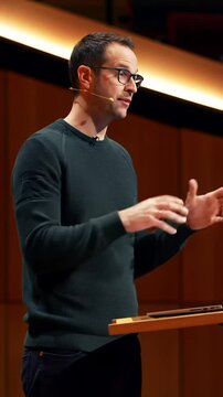 A charismatic mid-30s male speaker in a green merino wool sweater, wearing sleek glasses, gestures behind a modern podium with a glowing laptop, in razor-sharp focus with a blurred audience and
