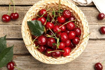 Fresh wet cherries in wicker basket and leaves on wooden table, flat lay