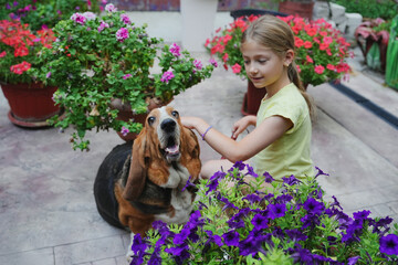 Girl sitting in garden with flowers and hugging basset hound