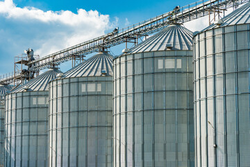 Metal silos are lined up in a storage facility under a clear blue sky with fluffy clouds,...