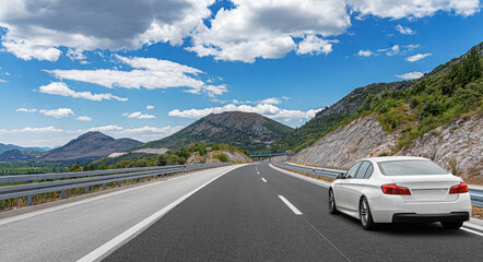 White car driving along a winding mountain highway under a vibrant blue sky, surrounded by lush...