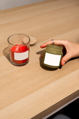 Two scented candles in stylish red and green jars with blank labels, placed on a wooden surface. A cozy mockup for holiday or home decor branding.