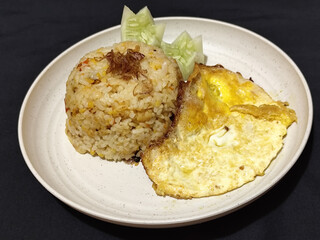 Fried rice served with sunny side up egg and fresh cucumber slices on a white plate, looks very delicious.
