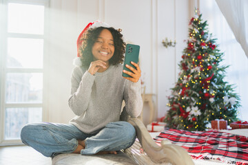 Merry Christmas. African woman in Santa hat having video chat in smartphone. Blogger in festive mood recording video in social network. Girl having video call with friends family celebrating Christmas