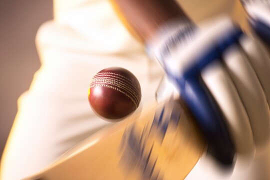 Dynamic close-up cricket batting action: professional player in white kit strikes red leather cricket ball with wooden bat wearing gloves.