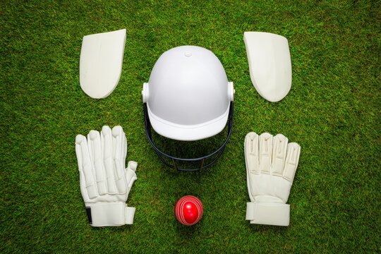 Premium cricket gear set: professional protective helmet, batting gloves, leg pads and red cricket ball on vibrant green turf background. - Powered by Adobe