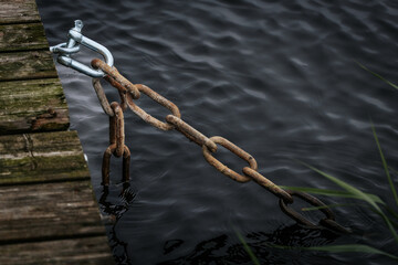 A weathered metal chain is attached to a wooden dock, partially submerged in dark rippling water,...
