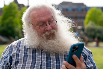 Elderly man with white beard reading a message on his smartphone in a park on a sunny day