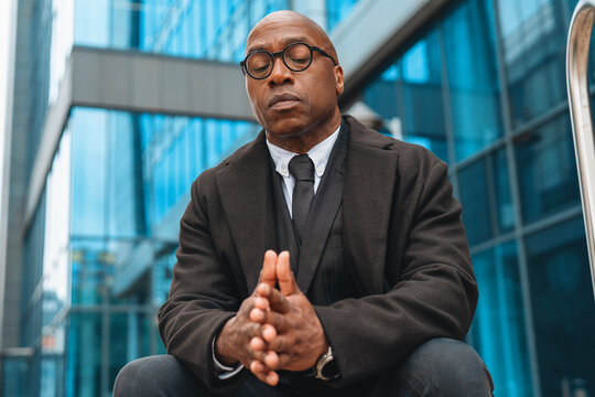 Middle-aged man in formal wear sitting pensively outside a modern building during daytime while wearing glasses and reflecting on thoughts