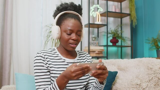 African American woman frustrated after fail losing video game on smartphone. Sitting on sofa in living room shaking head in irritation. Black girl looks disappointed, upset while playing game at home