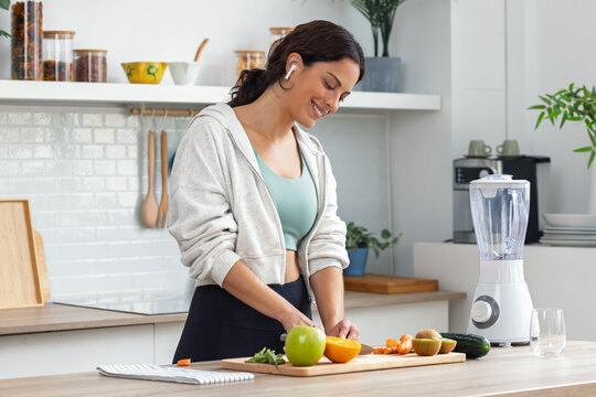 Athletic woman cutting fruits and vegetables to prepare a smoothie while listening to music with earphones in the kitchen - Powered by Adobe