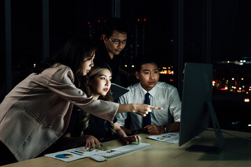 Group of Southeast Asian business professionals collaborating late night in office, woman pointing at glowing computer screen while colleagues analyze charts, symbolizing teamwork, dedication, 