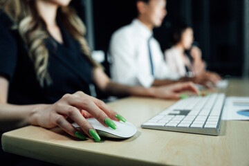 Female hand operating mouse at desk with coworkers on computers in background. Image highlights modern office environment and collaborative tech usage.

