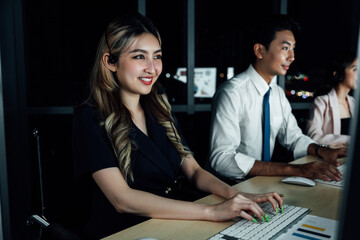 Smiling Asian businesswoman touches transparent virtual screen with stock graphs in background. Team analyzing financial data in modern office. Fintech and data interaction concept.