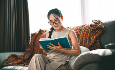 Beautiful young adult asian woman wearing eyeglasses reading book on sofa relax hobby routines
