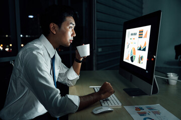 Asian businessman staying up late while drinking coffee and analyzing business charts on desktop...
