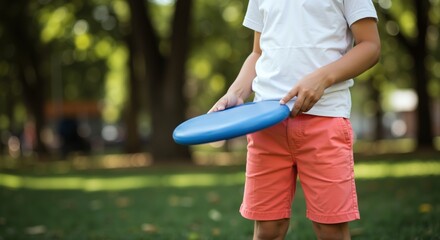 Boy in white shirt and coral shorts holding blue frisbee disc in green park outdoors. Child with sports equipment ready for active play. Summer recreation and youth fitness concept
