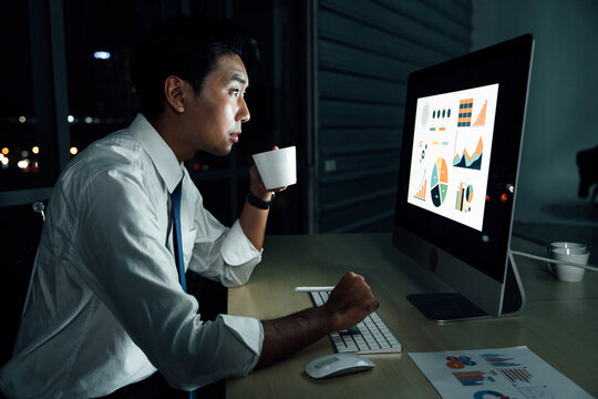 Asian businessman staying up late while drinking coffee and analyzing business charts on desktop screen. Night office illuminated only by monitor.