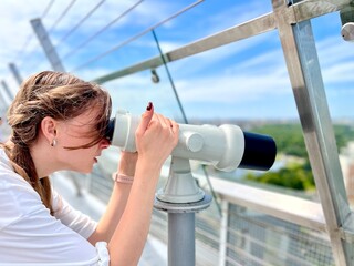 Profile view of a teenage young girl looking through stationary binoculars at cityscape from...