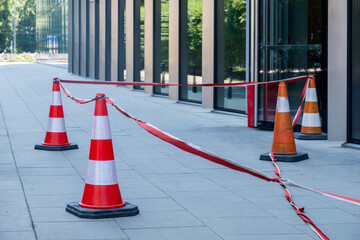 Traffic cones and caution tape are placed outside a modern building, indicating restricted access and safety measures in an urban environment with clear blue skies