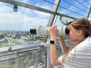 Young female tourist leans on public telescope while enjoying cityscape from observation platform....