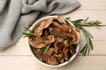 Pieces of dried mushrooms and rosemary in bowl on light wooden table, top view