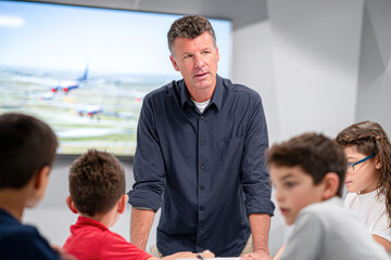 A man in a dark shirt stands at a table, engaging with four children in a classroom-like setting, with an airplane image displayed on a screen behind them.