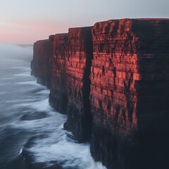Dramatic coastal cliffs at sunrise.  Ocean waves crash against towering, red-hued rock formations. Misty atmosphere