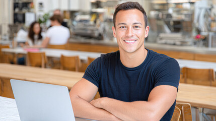 Fototapeta premium A young man sits at a table with a laptop, smiling confidently in a modern, casual work or study environment.