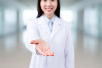 A smiling medical professional in a white coat extends an open hand forward in a bright, blurred clinical setting.