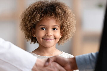 A young girl smiles warmly while two adults shake hands in the foreground, suggesting a positive and friendly interaction.