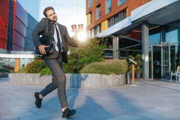 Young businessman rushing to office with coffee carrier, phone in hand