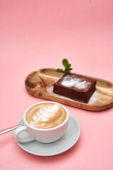Cappuccino with latte art and chocolate cake on wooden tray against pink background