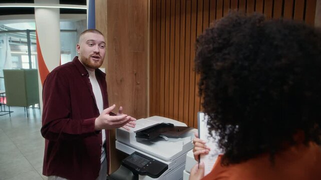 Stab shot of young woman with curly hair greeting male Caucasian colleague making copies of documents on office printer