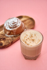 Cinnamon roll and latte on wood tray against pink background with sugar topping